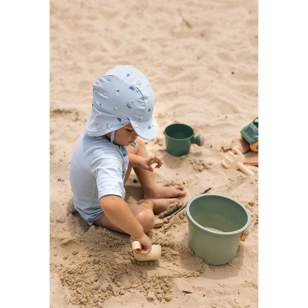 Criança com chapéu azul com desenhos de carros a brincar na praia com os brinquedos de praia